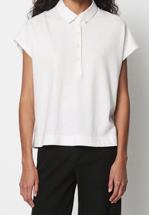 Frau trägt ein ärmelloses weißes Hemd mit Kragen und vier Knöpfen sowie schwarze Hose vor einem einfarbigen Hintergrund.