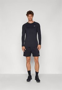 Άντρας που στέκεται φορώντας μαύρο αθλητικό μπλουζάκι με μακρύ μανίκι, μαύρες βερμούδες, μαύρες κάλτσες και μαύρα αθλητικά παπούτσια μπροστά σε απλό γκρι φόντο.