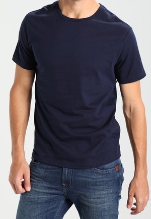 Homme portant un t-shirt bleu marine uni à manches courtes et un jean bleu, debout devant un fond clair uni.