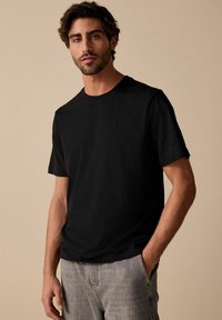 Camiseta negra de manga corta con cuello redondo, hecha de material suave. Lleva unos jeans gris claro y está de pie frente a un fondo neutro.
