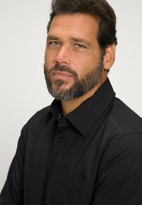 Camisa negra de botones con cuello, que presenta un bolsillo y una textura suave. La persona tiene barba y está mirando ligeramente de lado.