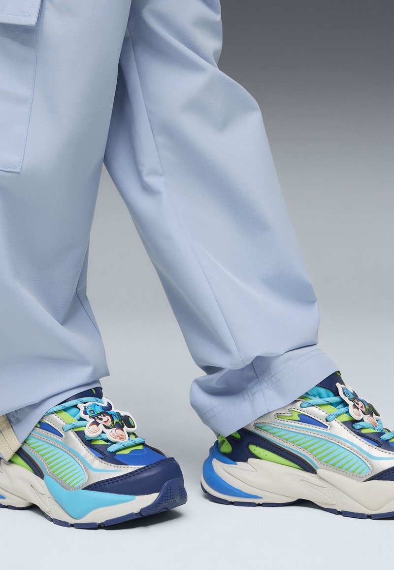 Chaussures de sport bleues, vertes et blanches avec des breloques de personnages de dessin animé sur les lacets, portées avec un pantalon bleu clair.