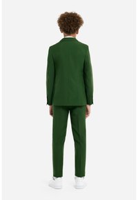 Completo verde con giacca su misura e pantaloni abbinati. Il tessuto ha una consistenza liscia. Indossato con sneaker bianche. Vista da dietro.