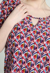 Personne ajustant un haut coloré à motifs avec des formes géométriques rouges, bleues, violettes et oranges et un petit lien au niveau de l'encolure en forme de trou de serrure.