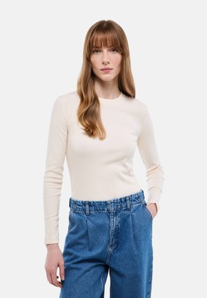 Jeune femme aux longs cheveux auburn portant un haut crème à manches longues et un jean ample en denim, sur un fond uni.