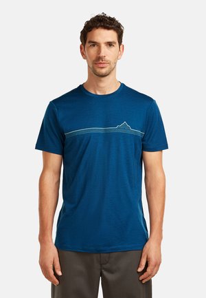Mann trägt ein blaues T-Shirt mit kurzen Ärmeln und einem minimalistischen Berglinien-Design über der Brust, steht vor einem einfarbigen Hintergrund.