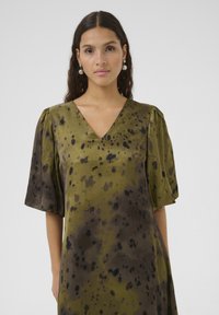 Maslina zelena satenska haljina s V-izrezom, kratkim napučenim rukavima i tie-dye uzorkom koji sadrži crne i tamnosmeđe mrlje.