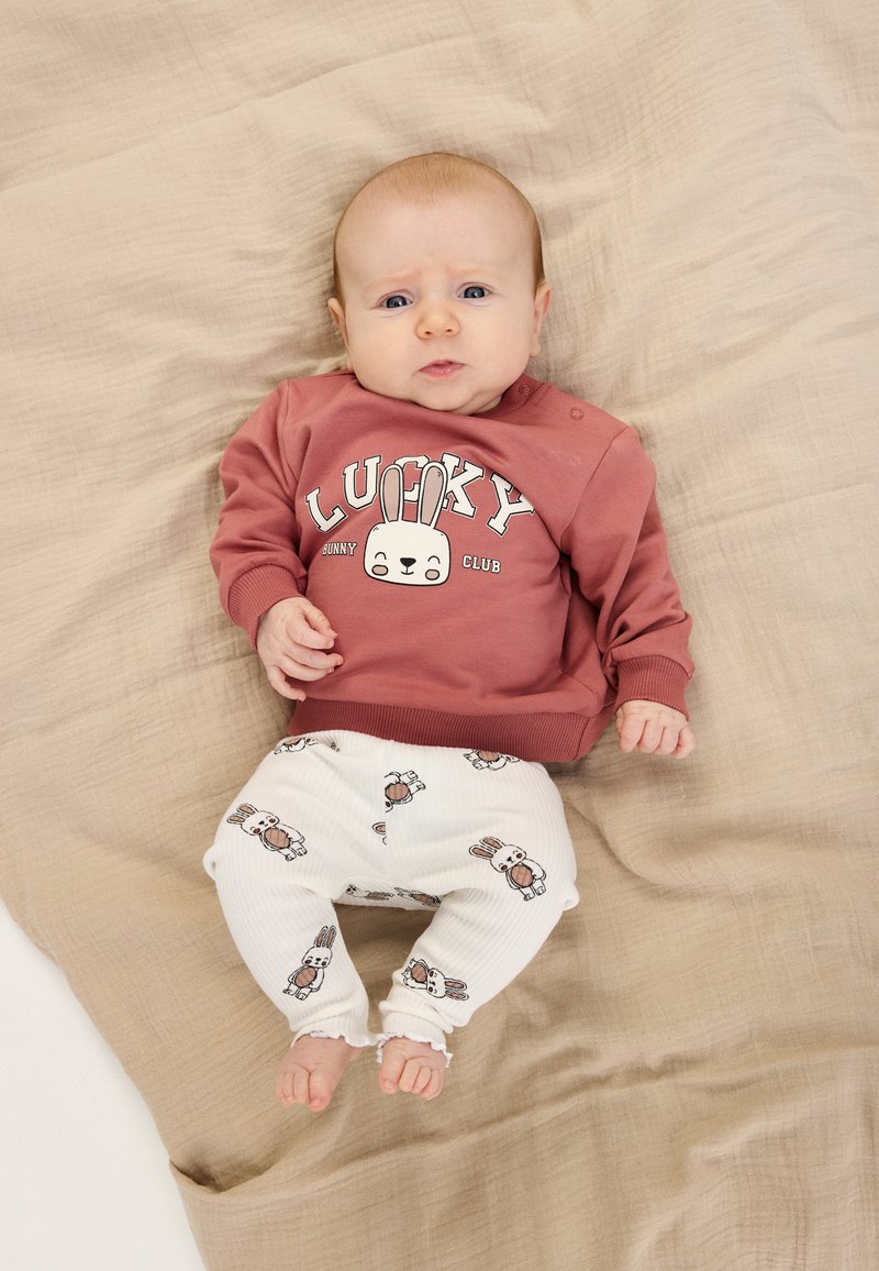Pink sweatshirt with "LUCKY" print and bunny design; white ribbed pants with bunny patterns; placed on a beige textured surface.