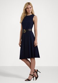 Vestido sem mangas azul-marinho com saia rodada, cintura ajustada por um cinto preto largo, acompanhado de saltos altos pretos com tiras. Textura de tecido suave.