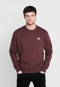 Bordeaux crewneck sweatshirt lavet af blødt materiale, med et lille hvidt logo på brystet og ribbede manchetter og kant.
