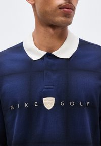 Άνδρας που φοράει μπλε σκούρο πόλο Nike Golf με λευκό γιακά και λογότυπο στο κέντρο του στήθους, περικομμένο στο λαιμό και στο μέσο του κορμού.