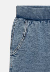 Hellblaue Denim-Jogginghosen mit geripptem Bund, ausgestattet mit orangen Nähten und einer seitlichen Taschenapplikation. Glatte Textur aus weichem Stoff.