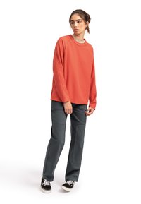 Felpa arancione ruggine con maniche lunghe e scollatura a girocollo, abbinata a pantaloni grigi scuri con toppe sulle ginocchia e sneakers nere.