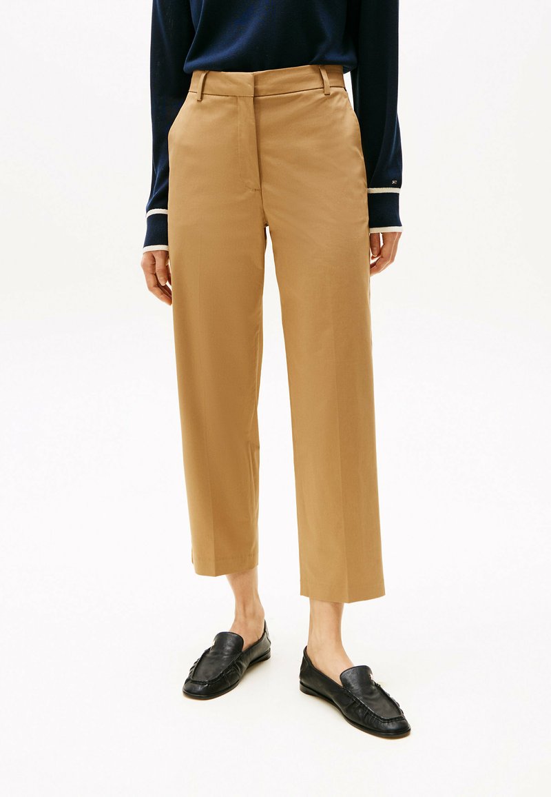Pantalons cropped taille haute beiges avec une texture lisse et un pli repassé, associés à des mocassins noirs. Comprend des poches avant et des passants de ceinture.