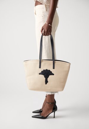 Femme portant un pantalon écru et des talons noirs, tenant un sac cabas beige avec des poignées noires et un emblème de fleur noire.