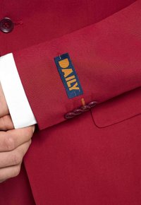 Blazer rosso con una texture liscia e una vestibilità sartoriale, con un risvolto della camicia bianca e un'etichetta blu con la scritta "DAILY" in lettere gialle.