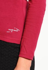 Långärmad röd topp i mjukt tyg, med rund halsringning och en vit "NAP NAF"-logotyp vid fållen. Matchas med svarta texturerade byxor.