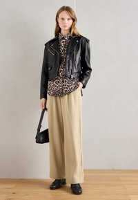 Woman stands against pale wall wearing black leather jacket, leopard print blouse, beige wide-leg pants, black shoes, and holding black handbag.