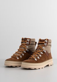 Brown suede hiking boots with beige textile upper, orange laces, silver eyelets, and rubber sole featuring a tread pattern.