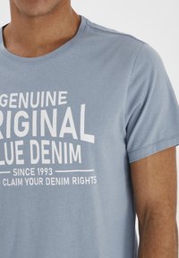 Lyseblå t-skjorte med korte ermer laget av bomull, med fet hvit tekst som leser "Genuine Original Blue Denim" med akcentlinjer under.