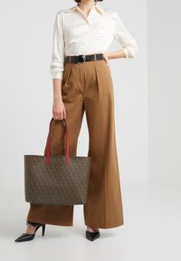 Brun mönstrad axelväska med röda handtag, bärs av en person i beige skjorta och bruna vida byxor, ihop med svarta spetsiga klackskor.