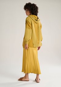 Sweatshirt à capuche jaune avec une coupe décontractée, design superposé et poignets élastiques, associé à une jupe longue assortie et des sandales marron.