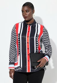 Blusa de botones con patrones geométricos audaces en negro, blanco y rojo. Mangas largas, cuello puntiagudo y combinada con un clutch negro.