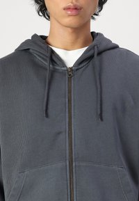 Graue Zip-Hoodie aus einem strukturierten Stoff, mit Kordelzugkapuze und Fronttaschen. Auffälliges Messing-Reißverschlussdetail in der Mitte vorne.