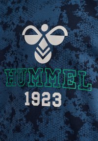 Marineblauwe stoffen met een patroon met daarop een wit bijenlogo boven "HUMMEL" in het groen en "1923" in het wit, met een hexagonale textuur achtergrond.