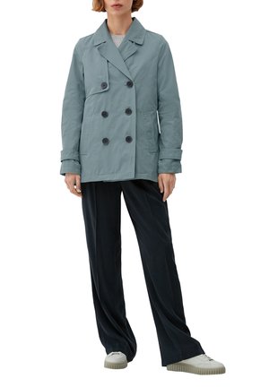 Veste double boutonnage bleu clair avec boutons noirs, poches latérales et col cranté, portée sur un pantalon large noir et des baskets blanches.