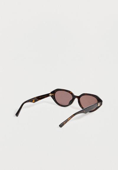 Lunettes de soleil à motif écaille avec verres ovales marron foncé et branches fines, posées sur une surface blanche unie.