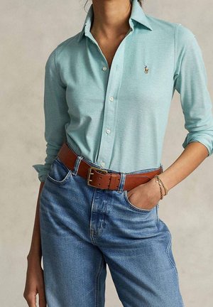 Vrouw die een lichtblauw overhemd met knopen draagt, ingestopt in een blauwe hoge taille jeans met een bruine leren riem, linkerhand in de zak, gouden armbanden om de pols.