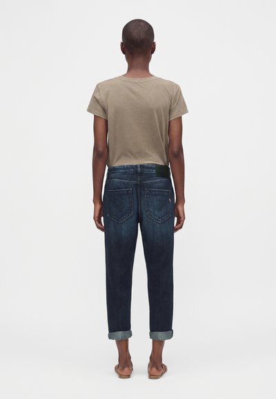 Personne debout de dos portant une chemise beige à manches courtes, un jean bleu foncé retroussé et des sandales marron sur un fond uni.