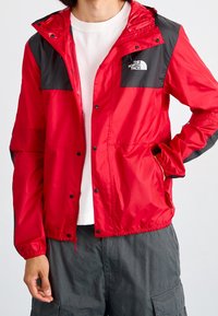 Czerwona wodoodporna kurtka z czarnymi akcentami, z pełnym zamkiem błyskawicznym, zatrzaskami i kapturem. Posiada logo na klatce piersiowej oraz elastyczne mankiety. Lekki materiał.