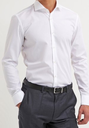 Mężczyzna ubrany w dopasowaną białą koszulę wizytową włożoną do ciemnoszarych eleganckich spodni z czarnym paskiem, z rękami w kieszeniach.