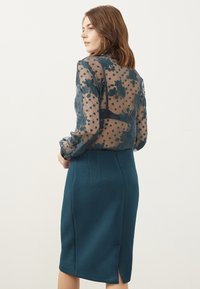 Transparante blouse met bloemen- en stippenpatronen, gecombineerd met een gestructureerde teal kokerrok met een rugsplit en geometrische ontwerpelementen.
