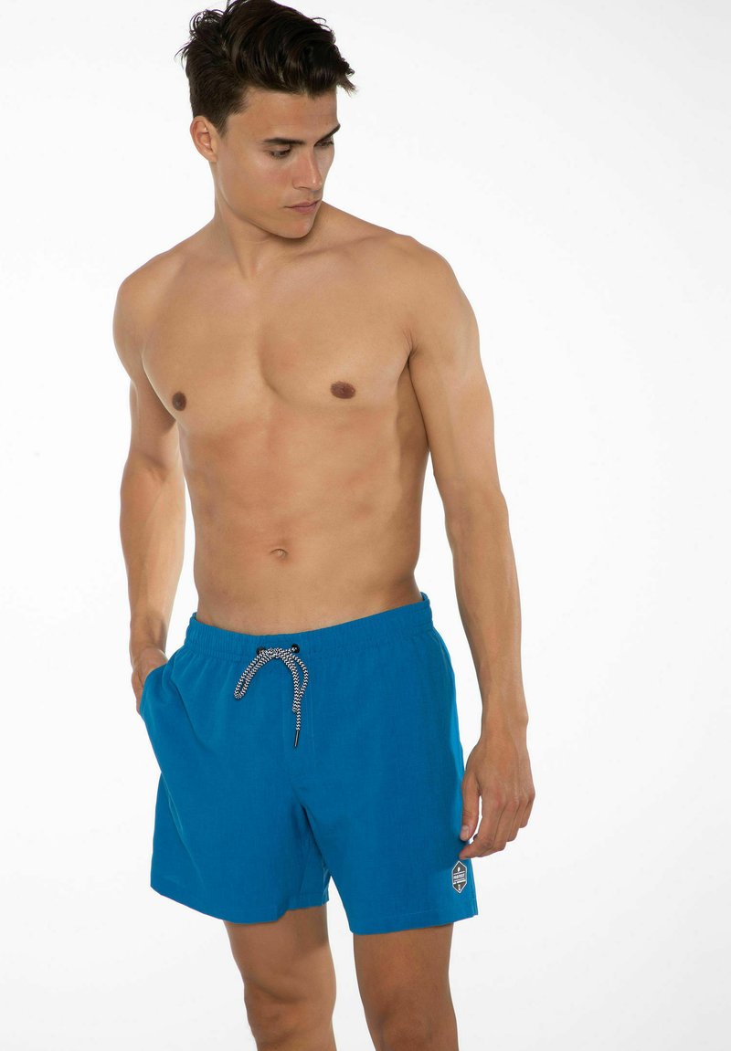 Blå simshorts med elastisk midjeband, justerbar dragsko i svart och vitt, samt en liten logotyp på vänster ben. Strukturerad väv.