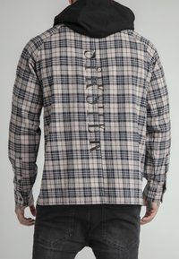 Camicia a quadri in beige e blu navy, realizzata in tessuto con finitura testurizzata. Presenta un cappuccio nero e testo stampato lungo la schiena.