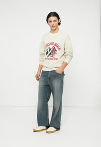 Sweter z grafiką "Jackson Hole, Wyoming" w czerwonym i zielonym kolorze na kremowym tle; połączony z jasnoniebieskimi szerokimi dżinsami i beżowymi sneakersami.