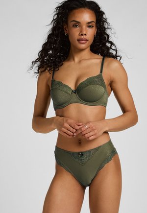 Vrouw met krullend haar die een bijpassende groene kanten beha en ondergoed draagt, staand met gevouwen handen voor zich, neutrale achtergrond.