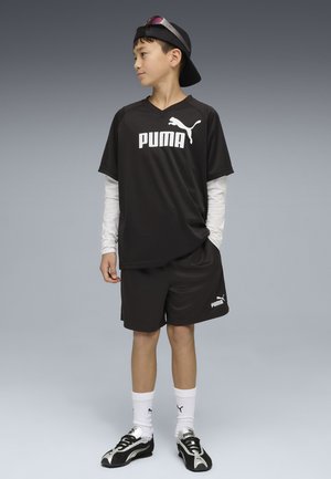 Ragazzo in piedi che indossa abbigliamento sportivo nero Puma, maglia a maniche lunghe bianca sotto la T-shirt, calzini bianchi Puma, scarpe da ginnastica nero e bianche e cappellino indossato all'indietro.