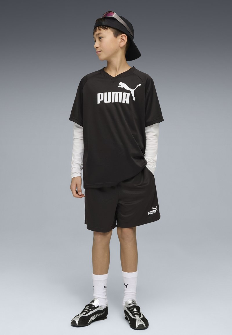 Garçon debout portant une tenue de sport noire Puma, un t-shirt à manches longues blanches sous le T-shirt, des chaussettes blanches Puma, des baskets noir et blanc, et une casquette portée à l'envers.