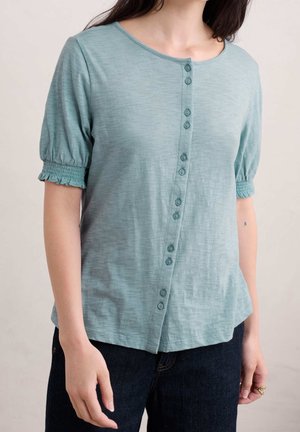 Kvinde iført en lyseblågrøn, kortærmet bluse med en diagonal række knapper og elastikkant på ærmerne, kombineret med mørke jeans.