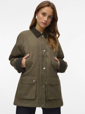 Žena oblečená v olivově zelené bundě s tmavým límcem a manžetami, s rukama v kapsách, čelící k přední části na bílém pozadí.