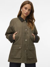 Kobieta ubrana w oliwkową kurtkę z ciemnym kołnierzem i mankietami, z rękami w kieszeniach, patrząca przed siebie na białym tle.