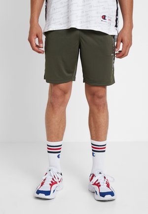 Hombre con pantalones cortos deportivos verde oliva, calcetines blancos hasta el tobillo con rayas rojas y azules, y zapatillas blancas, rojas y azules, de pie sobre un fondo liso.