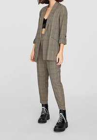 Costume à carreaux avec un blazer ample et un pantalon slim dans des tons de marron et de noir, associé à un haut en dentelle noire et des bottes noires épaisses.