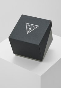 Svart sekskantet boks med teksturert overflate, med en sølvfarget triangelaktig Guess-logo på toppen, som hviler på en hvit pedestal.