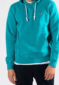 Turkos hoodie i mjukt material, med en kenguruficka, vita dragsnören och en liten logotyp på bröstet.