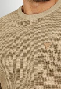 Maglione a girocollo in maglia beige testurizzata con piccolo logo in pelle a forma di triangolo sul petto, indossato da una persona che mostra collo e mento.